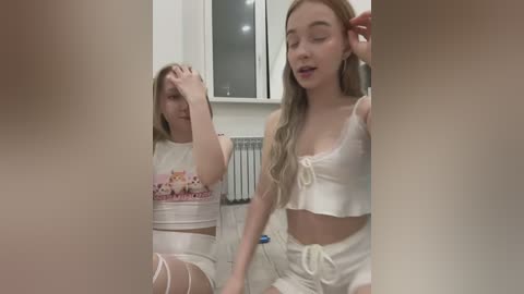 Media: Video of two young women in matching white crop tops and high-waisted shorts, one covering her face, the other smiling, in a modern, brightly lit room.