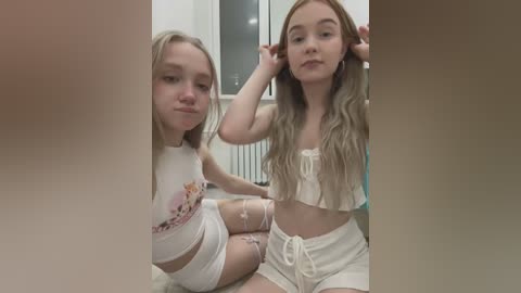 Media: Video of two young Caucasian girls with blonde hair, wearing white crop tops and shorts, sitting on a carpeted floor in a modern room.