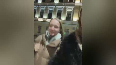 Media: Video of a young woman with light skin and shoulder-length blonde hair, wearing a beige coat and light blue scarf, standing in a large, dimly lit indoor space with high ceilings and columns.