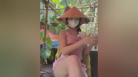 Media: Video of an Asian woman in a pink dress and conical hat, seated on a wooden bench in a lush, green garden with tropical plants, holding a glass of water.