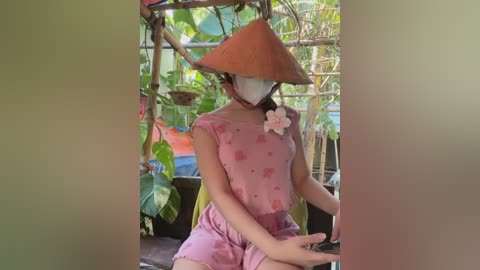 Media: Video of a person in traditional Vietnamese conical hat, pink floral dress, and face mask, seated in a green, leafy garden.