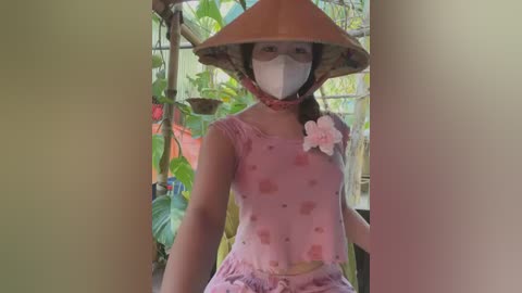 Media: Video of an Asian woman in a pink floral dress, wearing a traditional conical hat, mask, and floral brooch, standing amidst lush greenery and hanging plants in a garden.