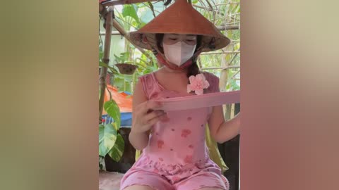 Media: Video of a young woman with light skin, wearing a pink dress with floral patterns, a wide-brimmed hat, and a face mask, reading a pink book in a lush green garden.