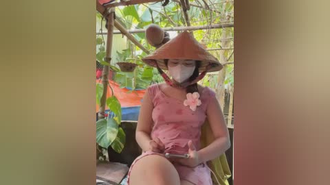 Media: A video of an Asian woman in a pink dress, wearing a conical hat, sitting in a bamboo hut surrounded by greenery. She's holding a smartphone.