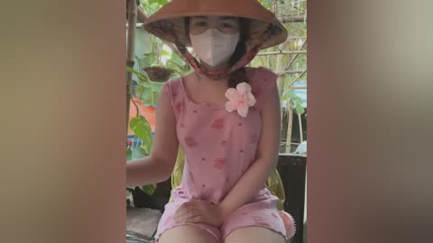 Media: Video of a young Asian girl with light skin, wearing a pink floral dress, a white mask, and a conical hat adorned with a pink flower, sitting on a chair in a lush garden.