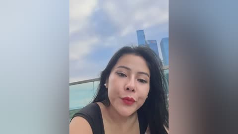 Media: Video of a young Asian woman with long black hair, red lipstick, and a black top, smiling from behind a glass wall, with a cloudy sky and cityscape visible in the background.