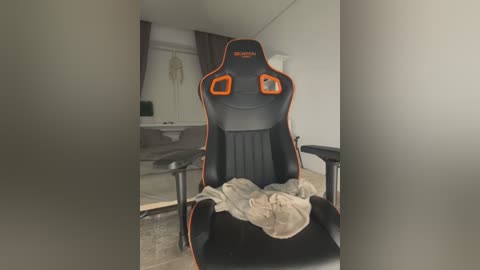 Media: Video of a black gaming chair with orange accents, covered in a beige blanket, in a cluttered room with a desk, computer monitor, and wall art.