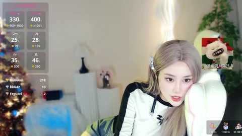 Media: A video of a young Asian woman with long blonde hair, wearing a black and white hoodie, resting her head on a white pillow, in a cozy, festive room with a Christmas tree and a green plant in the background.