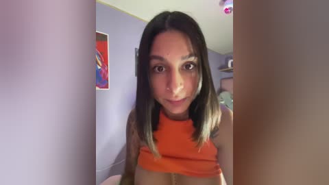 Video of a young woman with medium skin tone and long, straight black hair, wearing an orange crop top. Background features a purple wall with colorful artwork and a bed.