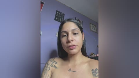 Media: Video of a topless Latina woman with medium-brown skin, long black hair, and multiple tattoos, including a heart on her chest, standing in a purple-painted room with wall decorations.