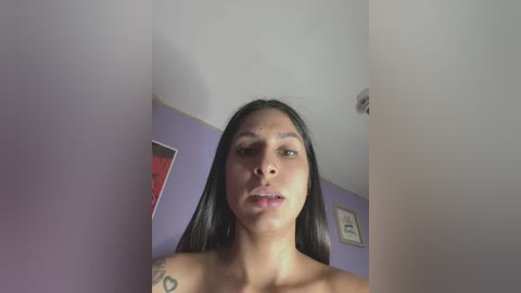 Media: Video of a topless woman with long, straight, dark hair, light brown skin, and a tattoo on her shoulder, standing in a room with purple walls and framed art.