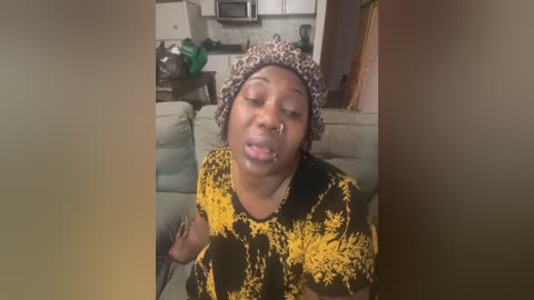 Media: Video of a middle-aged woman with dark skin and a nose ring, wearing a black and yellow patterned dress, and a leopard-print headscarf, seated on a couch in a dimly lit living room with a kitchen backdrop.