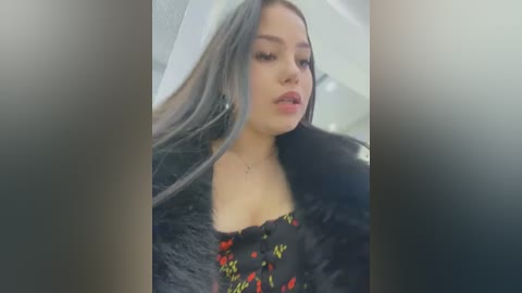 Media: Video of a young woman with long, platinum blonde hair, wearing a black fur coat with a floral-patterned dress underneath. Her skin is fair, and she has a neutral expression. The background is blurred.