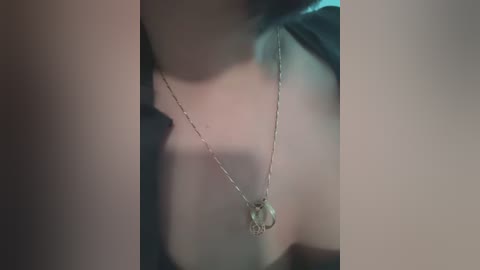 Media: A close-up video of a delicate silver necklace with a tiny, intricate pendant, worn by a person with fair skin, wearing a dark green top. The background is blurred, emphasizing the necklace.