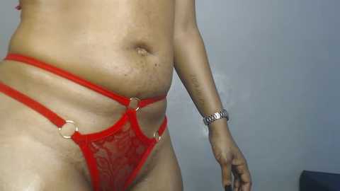 Media: Video of a person with medium brown skin, wearing a red lace thong with gold rings, and a silver wristwatch, standing against a plain gray wall.