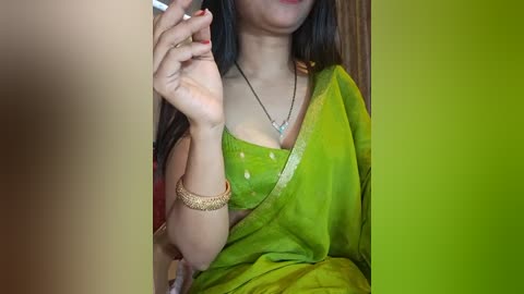 Media: Video of a light-skinned woman with long black hair, wearing a green sari with gold accents, gold bangles, and a necklace, holding a cigarette in her right hand, indoors with a green gradient background.