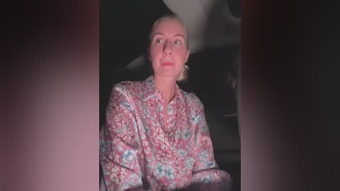 Media: Video of a middle-aged white woman with blonde hair, wearing a floral-patterned blouse, gazing upwards, possibly in a car, with a dark, dimly lit background.