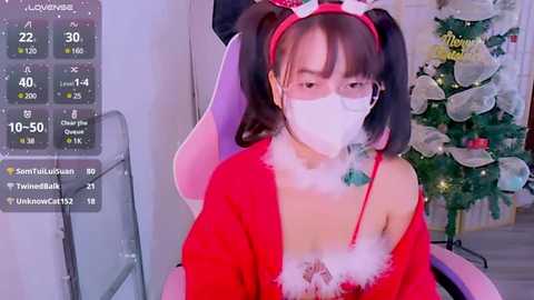 Media: Video of an Asian woman with a slender build, wearing a red Santa costume with white fur trim, and a face mask. She sits in front of a decorated Christmas tree.