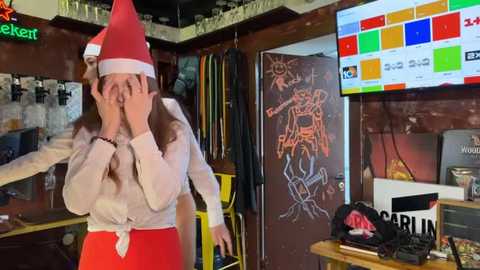 Media: Video of a woman in a red Santa hat, hiding her face with hands, wearing a white blouse and red skirt in a cozy, dimly lit bar with graffiti, a large TV, and bar stools.