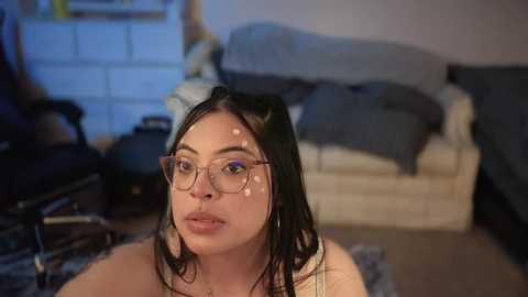 Media: Video of a young woman with long black hair, fair skin, and large round glasses, sitting on a plush carpet in a cozy living room. She has a neutral expression, and the room features a beige couch, dark furniture, and a white brick wall.