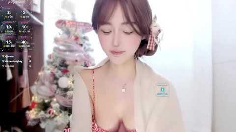 Media: Video of a young Asian woman with fair skin, brown hair, and a red polka-dot bra, wearing a beige cardigan, sitting next to a decorated Christmas tree.