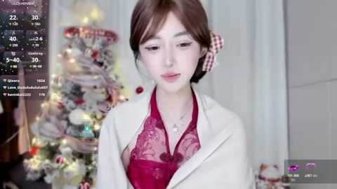 Media: Video of a young Asian woman with light skin and brown hair, wearing a red lace blouse under a white cardigan, standing in front of a decorated Christmas tree.