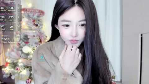 Media: A video of a young East Asian woman with long, straight black hair, fair skin, and a soft smile, wearing a beige fur-trimmed coat, standing in front of a Christmas tree with colorful ornaments.