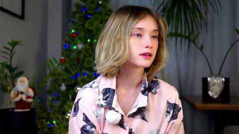 Media: Video of a young girl with shoulder-length blonde hair, wearing a pink shirt with black cat faces, standing in a softly lit room with a Christmas tree, potted plants, and festive lights.