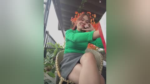 Media: Video of a smiling, middle-aged Asian woman with short red hair, wearing a bright green top and gray skirt, sitting on a wicker chair on a balcony with lush green plants.