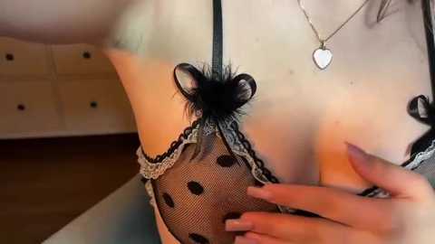 A video of a woman in black lingerie with polka dots, black lace, and a heart pendant. Her hand is touching her breast. Background includes a dresser.