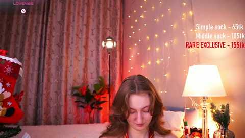 Media: A video of a woman with light brown hair, wearing a festive red dress, sitting on a bed adorned with holiday decorations. The room is warmly lit with string lights and a white lamp.