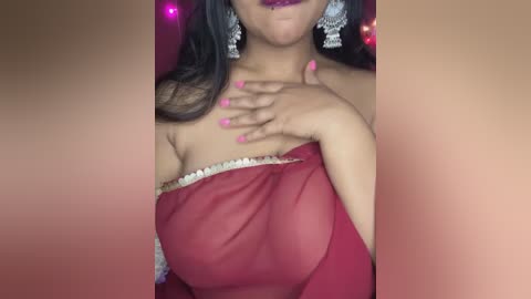 Media: Video of a woman with medium skin tone and long dark hair, wearing a sheer red off-shoulder top with white lace trim. She has pink nail polish and silver earrings, posing against a pink background with blurred lights.