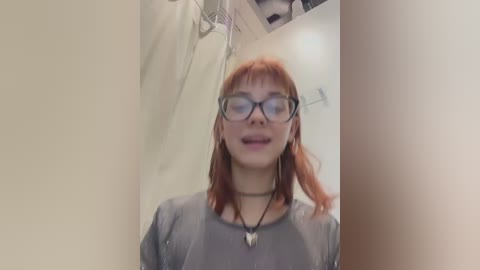 Media: Video of a young woman with long red hair, wearing black-rimmed glasses and a grey shirt, standing in a shower with clear glass door.