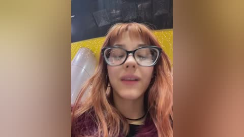 Media: Video of a young woman with long, reddish-brown hair and large, black-rimmed glasses, wearing a black and maroon striped shirt. Background features a yellow and black patterned cushion.