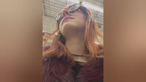 Media: Video of a fair-skinned woman with long, reddish-brown hair, wearing large, black-framed glasses and a dark fur coat, standing in a warehouse with exposed beams and industrial lighting.