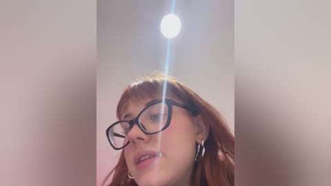 Media: Video of a fair-skinned woman with long, reddish-brown hair, wearing black-framed glasses and large silver hoop earrings, smiling, with a bright, blurry light source in the background.