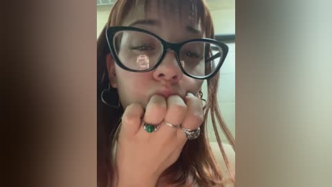 Media: Video of a young woman with fair skin, long auburn hair, wearing large black-framed glasses, and kissing her fingers. She's adorned with a green ring and silver bracelet. Background is blurred, possibly indoor.