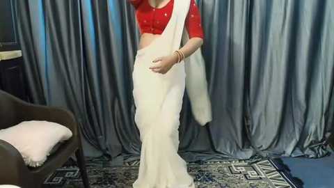 Media: Video of a woman in a red blouse and white sari, standing in front of grey curtains, with a brown armchair and patterned rug in the background.