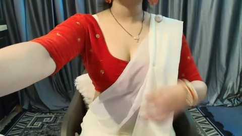 Media: A video of a fair-skinned woman with long dark hair, wearing a red blouse with a white sari, seated indoors with gray curtains and geometric-patterned floor.