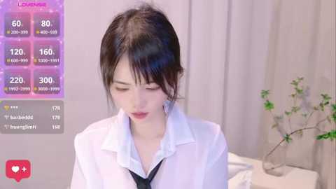 Media: Video of an East Asian woman with fair skin and dark hair in a white shirt and black tie, sitting in a modern room with a plant.