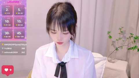 Media: Video of a young Asian woman with dark hair, wearing a white shirt and black tie, in a softly lit bedroom with a green plant.