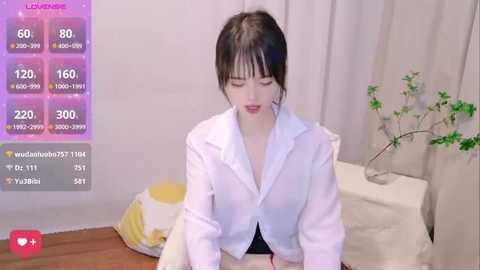 Media: Video of a young Asian woman with pale skin and dark hair, wearing a white hoodie, kneeling on a wooden floor in a minimalist room with a plant and white curtains.