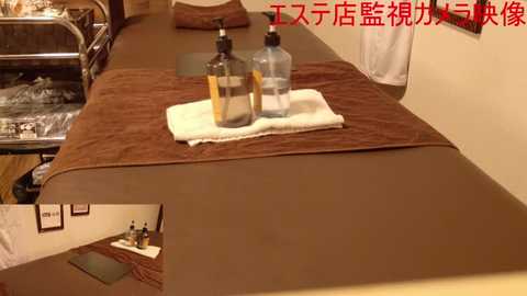 Media: Video of a spa treatment room with a wooden table covered in a brown towel. Two clear bottles of massage oil sit on a folded white towel. The room features beige walls and a metal table with a mirror.