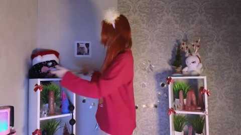 Video of a woman in a red jacket and headband, adjusting a Christmas wreath on a white shelving unit with festive decorations, against a wallpapered wall with a framed photo.