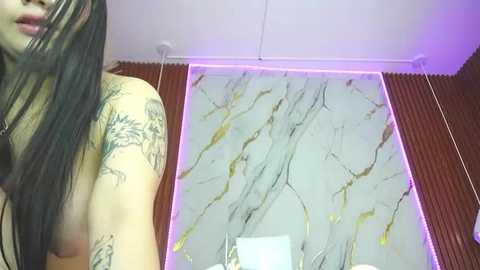 Media: Video of a topless woman with long black hair, showing tattoos on her left arm, standing in a room with a marble-effect glass shower door and red-brown wooden paneling.