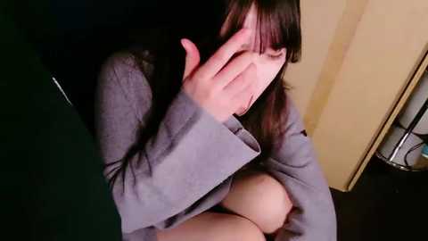 Media: A video of an Asian woman with long black hair, wearing a gray sweater, sitting on a dark wooden floor, covering her face with her hand, in a dimly lit room with beige walls and a metal shelf in the background.