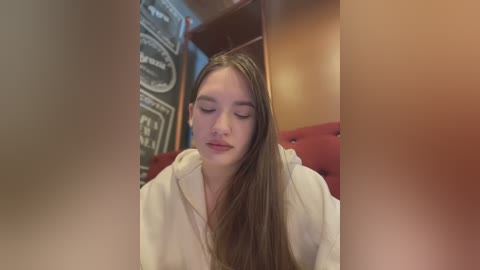 Media: Video of a young, fair-skinned woman with long, straight brown hair, wearing a white robe, sitting in a dimly lit room with a chalkboard wall and red cushioned seats.