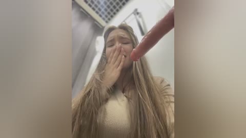 Media: A video shows a blonde woman with long hair, wearing a beige sweater, covering her mouth with her hand. A hand, presumably male, with a long fingernail, is close to her face. The background is a tiled bathroom.