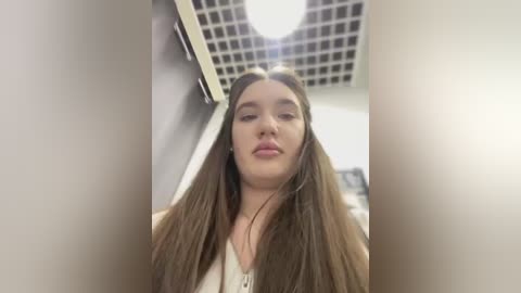 Media: Video of a young Caucasian woman with long brown hair, fair skin, and full lips, wearing a beige top, standing indoors against a light-colored wall with a grid-patterned ceiling.