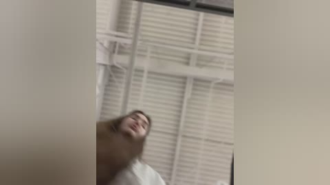 Media: A blurry video of a person, possibly a woman, with light skin, lying on a bed with white blinds. The room has white walls and a ceiling fan, creating a clinical, minimalist atmosphere.